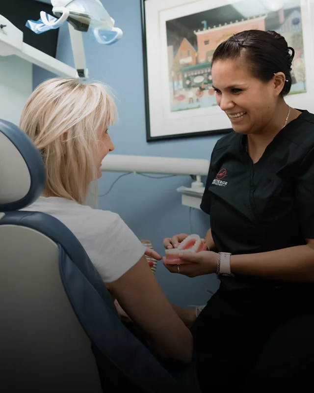 A patient sitting in an exam chair and pointing at a tooth model held by a hygienist